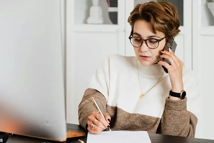 Frau in stilvoller Kleidung spricht am Handy und macht Notizen in einem hellen Büro.