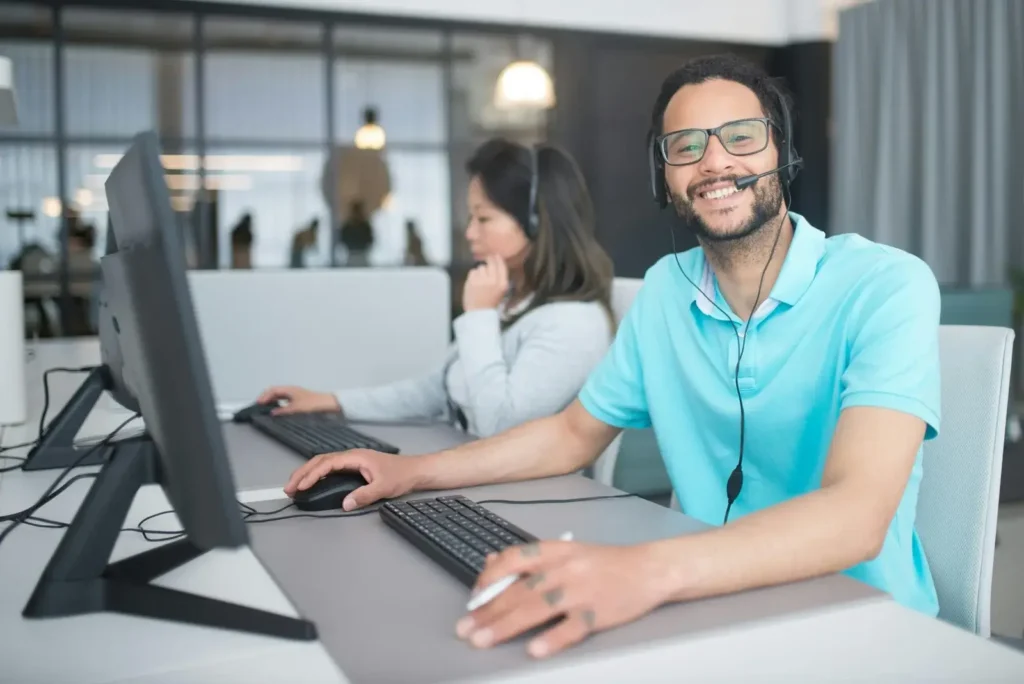 Freundlicher Kundenbetreuer mit Headset arbeitet am Computer in einem hellen Großraumbüro.