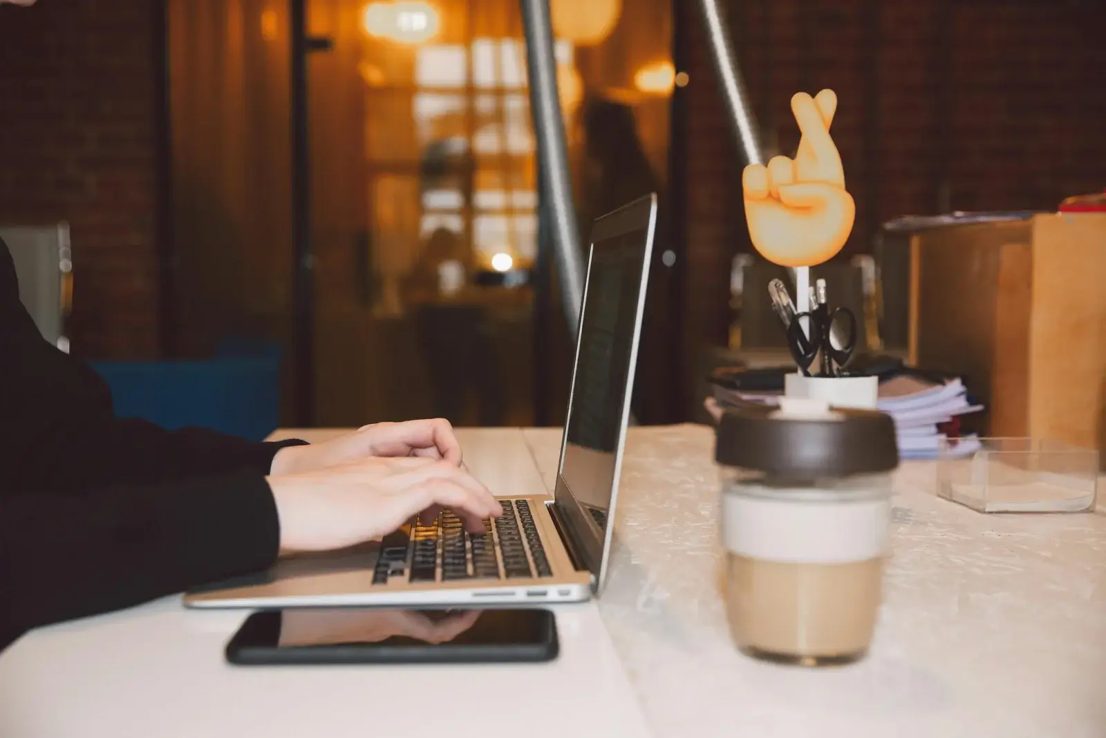 Person tippt auf einem Laptop in einem gemütlichen Café-Arbeitsplatz mit stimmungsvoller Beleuchtung und einer Tasse Kaffee im Vordergrund.